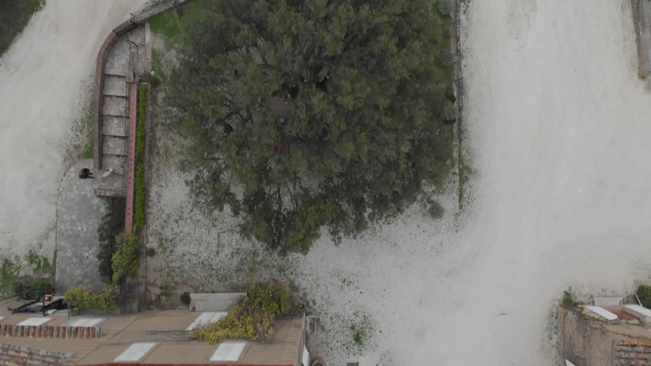 Aerial top-down dolly-forward shot over rustic rooftops as a person exits one door and walks across a courtyard to another entrance
