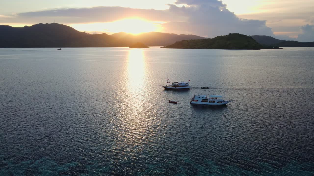 Drone footage of boats sailing under golden sunrise rays in the Komodo National Park sea, Indonesia. Gliding on the deep dark blue sea with the silhouette of surrounding islands as the backdrop.