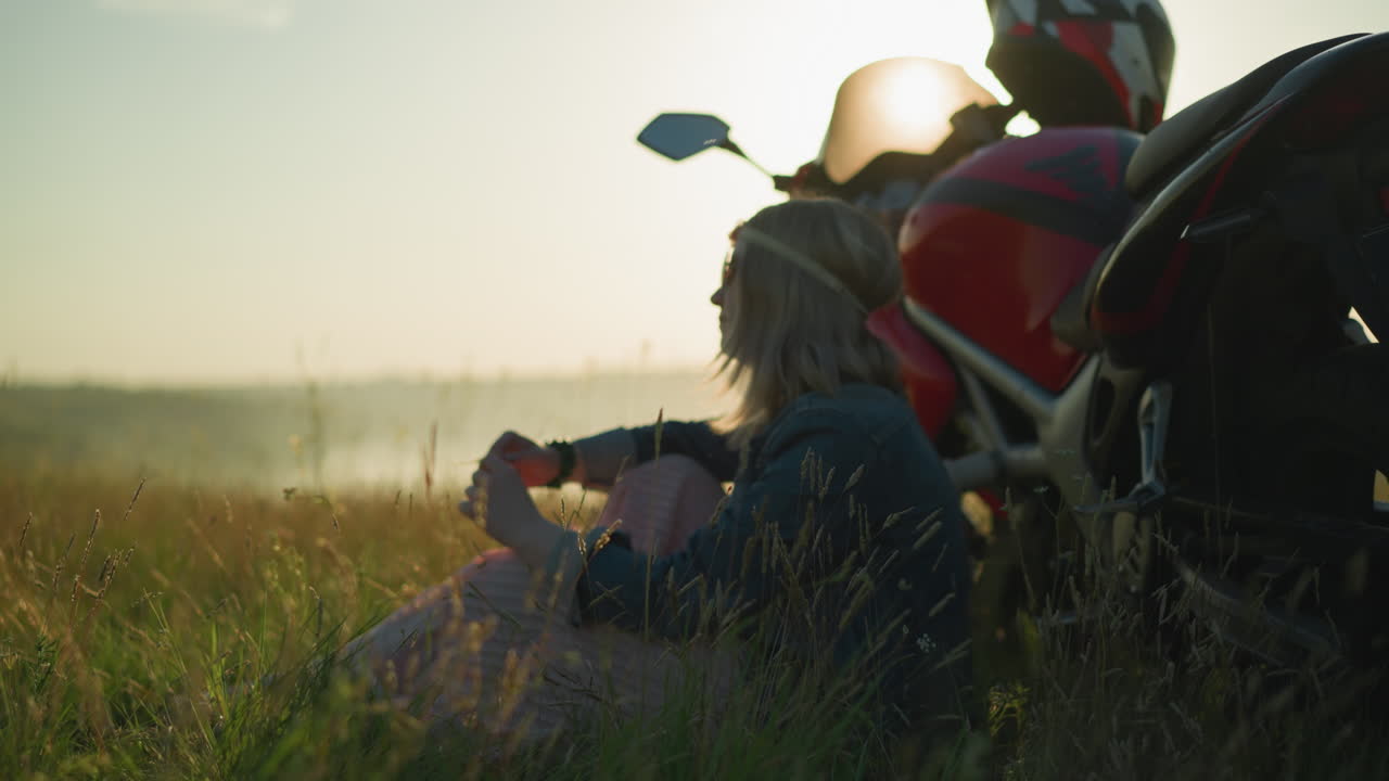 A woman with a flower headband and sunglasses sits in a grassy field beside a motorcycle, she thoughtfully plays with a strand of grass in her hand, lost in thought, as the sun sets behind her
