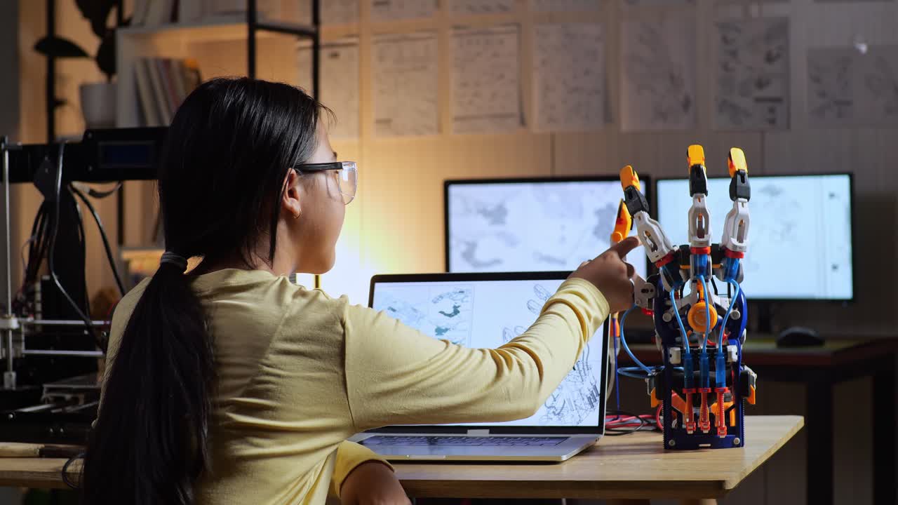 vista trasera de una adolescente asiática con impresión 3d comprobando una mano de cyborg mientras la diseña en una computadora portátil en casa