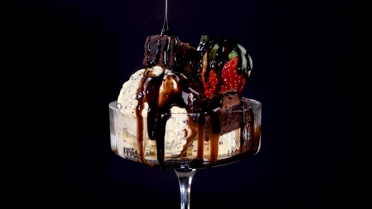 Chocolate sauce streams onto vanilla ice cream topped with strawberries, brownie pieces, and chocolate chips in a glass dish, under dramatic studio lighting and dark background