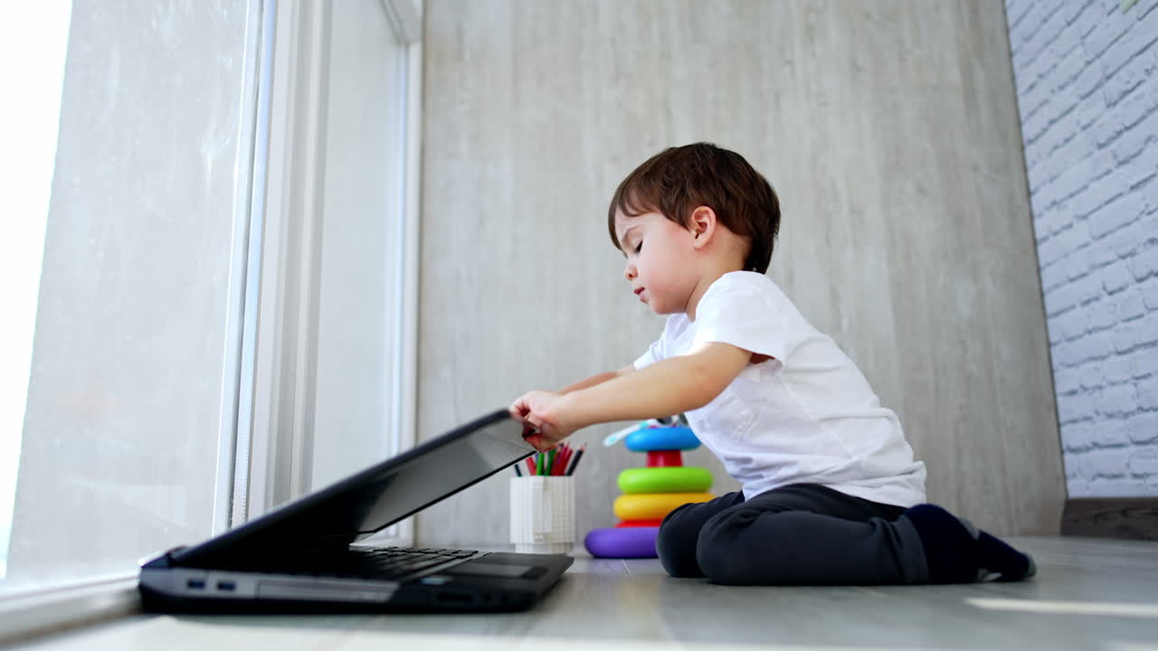 Cute toddler sitting on the floor opens the laptop. Baby boy presses on keys on the keyboard one by one. Low angle view.