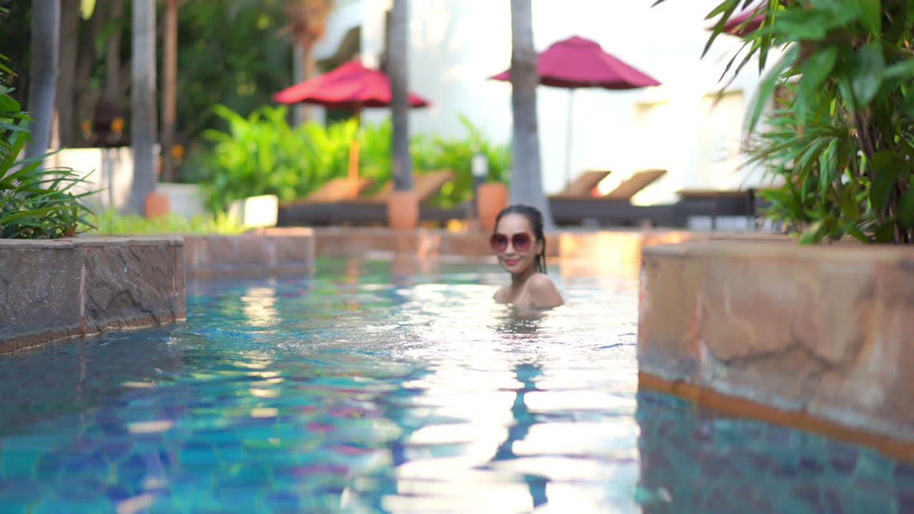 A young woman wearing sunglasses wades into a resort swimming pool slowly turns to interact with the camera