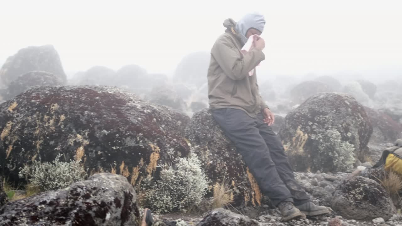 A black man rests and blows his nose with a backpack and jacket on a boulder during fog