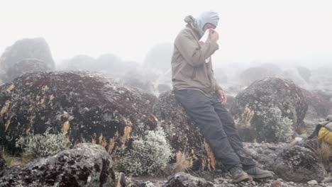 a black man rests and blows his nose with a backpack and jacket on a boulder during fog