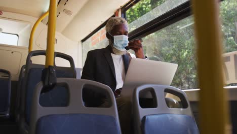 Afroamerikanischer-älterer-Mann-Mit-Gesichtsmaske,-Der-Im-Sitzen-Auf-Dem-Smartphone-Spricht-Und-Einen-Laptop-Benutzt