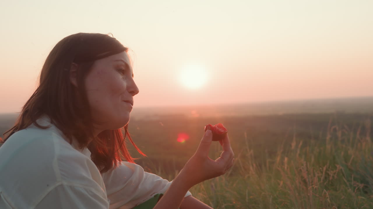 Side view of elegant lady in white shirt biting into juicy ripe plum while sunset light flares across her profile, seated in tall grassy meadow with blurred countryside