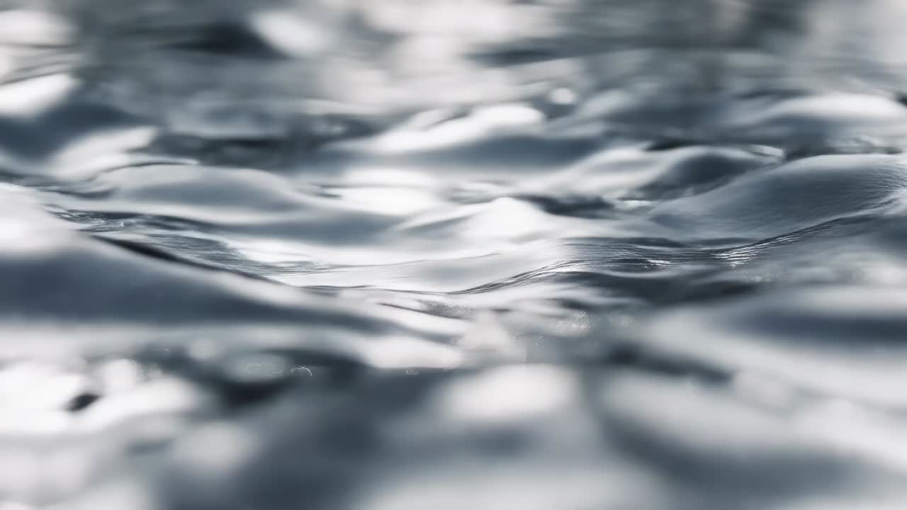 Superficie increspata dell'acqua a livello del pelo dell'acqua, innescata da goccioline che attraversano l'inquadratura formando riflessi