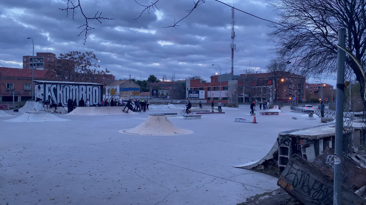 Giovani che praticano sport d'azione e cultura di strada in uno skate park di cemento con rampe e graffiti, sotto un cielo coperto.