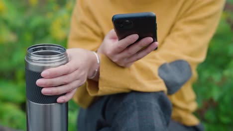 Young-woman-outdoors-browsing-on-her-phone-and-holding-a-steaming-thermos