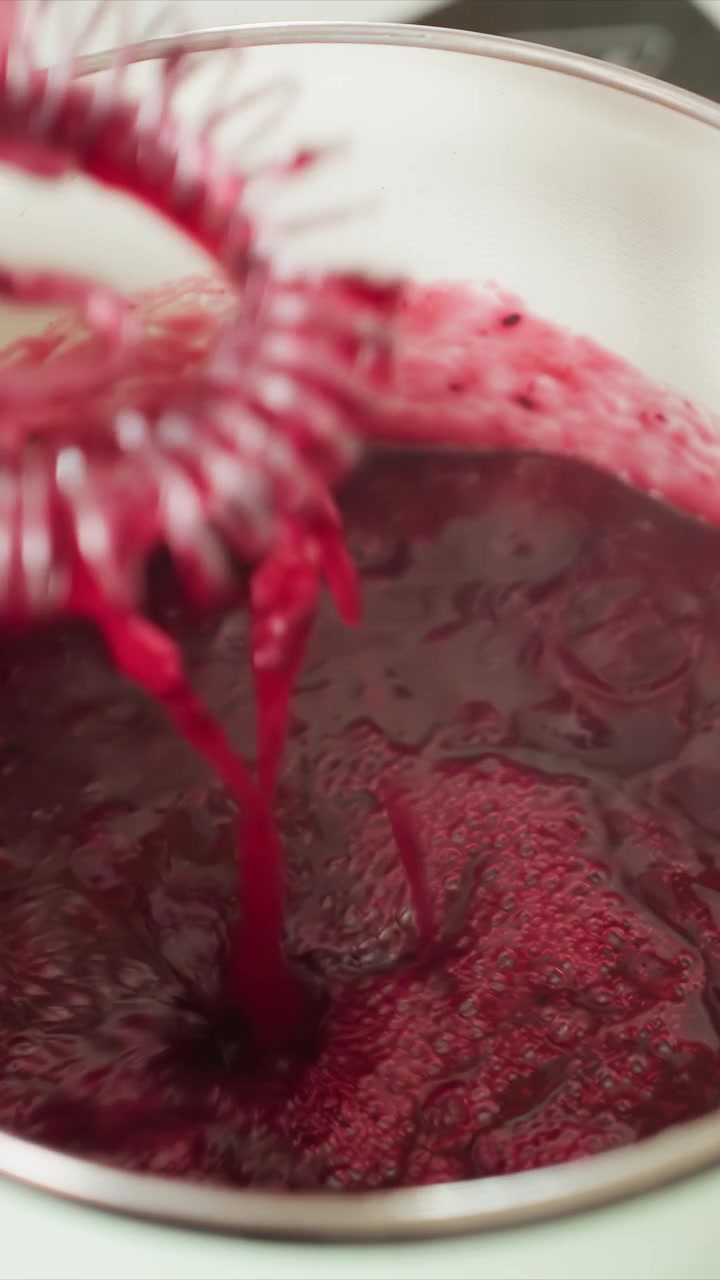 Close up of thickening red liquid being stirred in white container. Small black seeds are. Scene is set in kitchen with minimal background details focusing with stirring