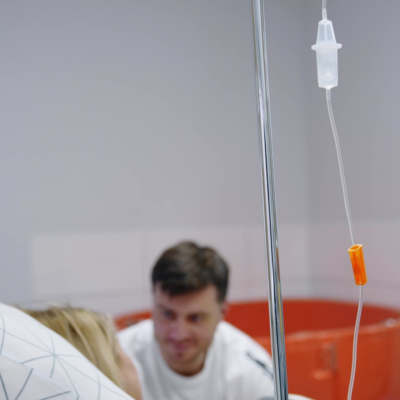 Drop counter near the bed of Caucasian woman. Man talks to his pregnant wife in hospital ward at backdrop in blur