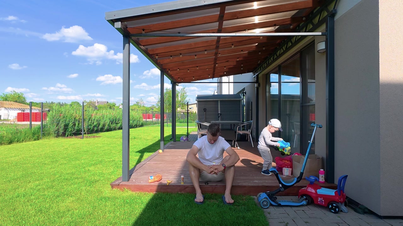 Otec relaxuje bosýma nohama na dřevěné terase, zatímco si jeho malé dítě hraje s hračkami a koloběžkou na sluncem zalité zahradě. Moderní dům, svěží zelený trávník, jasné denní světlo, radostný rodinný okamžik.
