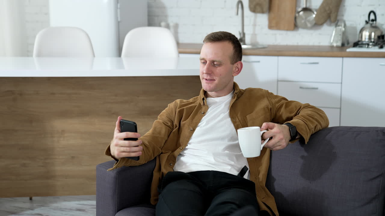 Handsome man using a phone at home. Guy rests on sofa with a cup of coffee and doing photos on a smart phone in the kitchen.