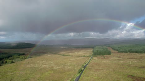 High-aerial-drone-footage-in-the-rain-rolling-left-above-a-long,-straight-road-looking-towards-a-birght-rainbow-with-dark-grey-clouds-in-the-background,-with-fields,-trees-and-moorland-in-Scotland