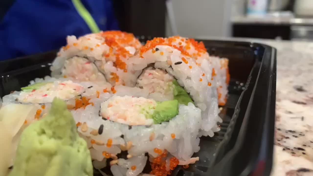 Child picking up a sushi roll from a take out container. California rolls with bright orange masago eggs and black sesame seed. Wasabi and ginger on the side.
