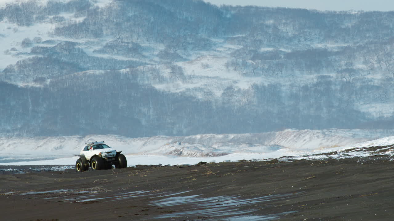 Terrænkøretøj kørende på sort sandstrand med sneklædte bjerge, store stranddæk med skovklædte klipper, Kamchatka, Rusland, slowmotion.