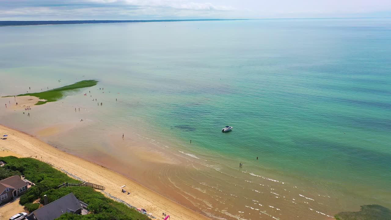 Sweeping aerial footage of a coastal flat at low tide reveals sculpted sandbars, reflective tide pools, a beached boat, and colorful patches of marsh grass set against the open shoreline