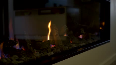 Slow-motion-close-up-of-gas-indoor-fireplace-with-hot-flames-embers-artificial-wooden-logs-behind-glass-panel-household-heating-fuel-modern-home-design