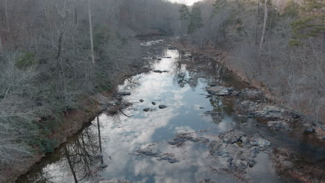 Stagnant-Rocky-River-Reflecting-Cloudy-Sky-In-Ominous-Forest-In-Fall