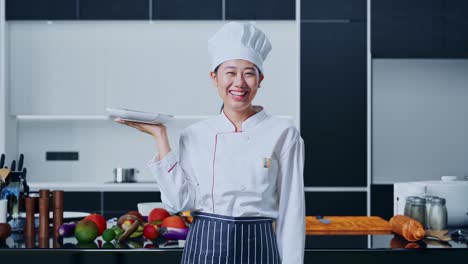 mujer chef asiática sosteniendo un plato y sonriendo a la cámara en la cocina de casa