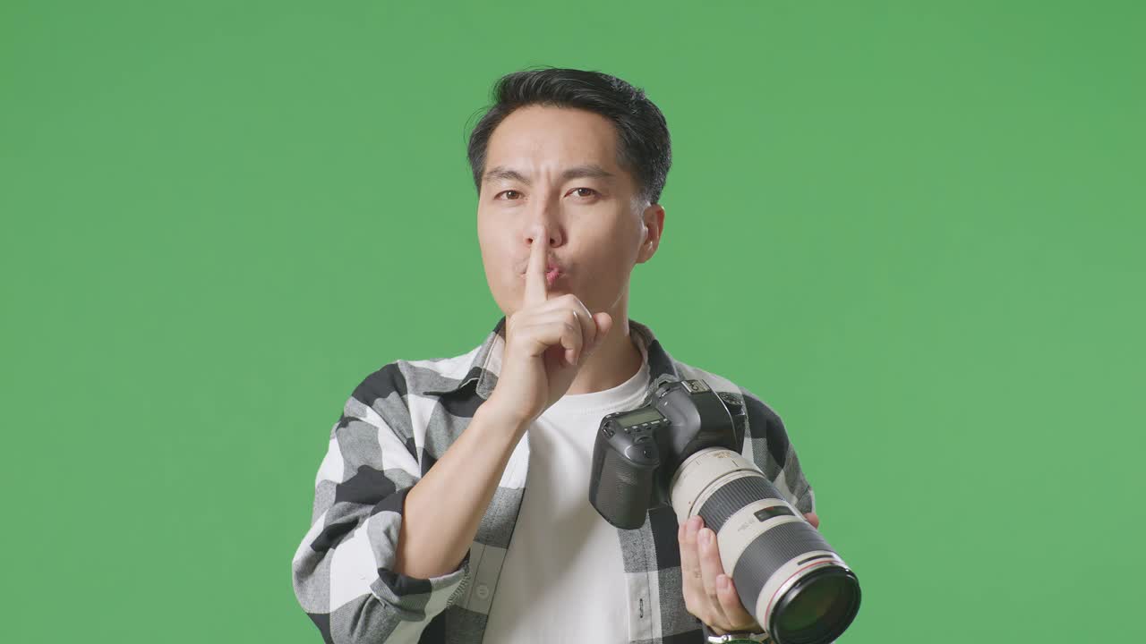 스튜디오에서 녹색 화면 배경에 서서 사진을 찍고 shh 제스처를 하는 카메라를 사용하는 아시아 사진작가의 클로즈업