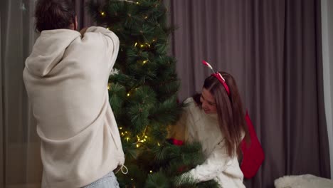 A-brunette-guy-in-a-white-sweatshirt-together-with-his-brunette-girlfriend-in-a-white-sweater-decorate-the-Christmas-tree-and-prepare-for-the-Christmas-holiday-in-their-cozy-home-in-winter.-Happy-loving-couple-guy-and-girl-preparing-for-Christmas-and-decorating-the-Christmas-tree-with-bright-garland