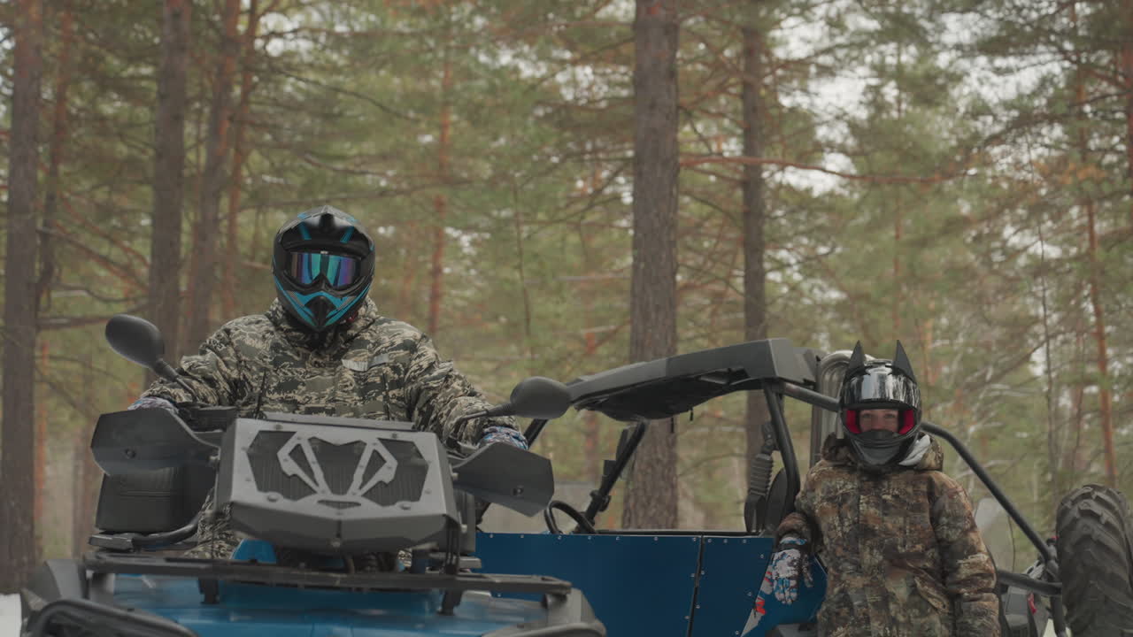 Pilota mimetizzato su ATV in una foresta innevata, prepara il quad con casco e occhiali. Un compagno è accanto al veicolo, il motore è al minimo. Tronchi di pino e terreno innevato creano un'atmosfera di avventura invernale selvaggia.