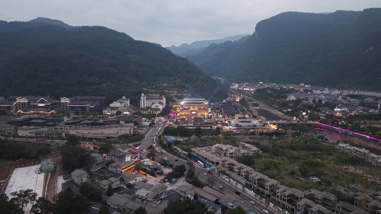 Lenta panoramica aerea su un villaggio fluviale di Zhangjiajie al crepuscolo, con un ponte illuminato di blu ed edifici illuminati lungo il corso d'acqua, tetti fitti sottostanti e montagne carsiche sullo sfondo, Wulingyuan, Zhangjiajie