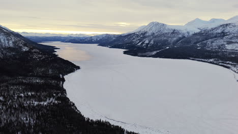 frozen kusawa lake through mountains at sunset in winter in yukon, canada. aerial shot