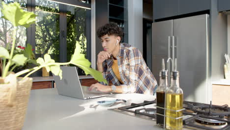 using laptop in kitchen, teenage boy with earbuds focusing on work