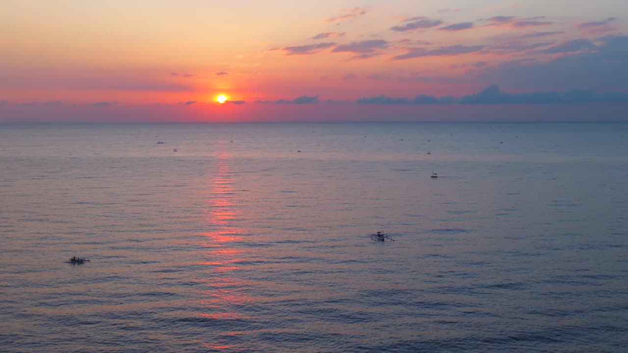 Aerial sunrise over pastel sea dotted with small fishing boats and a pink orange horizon