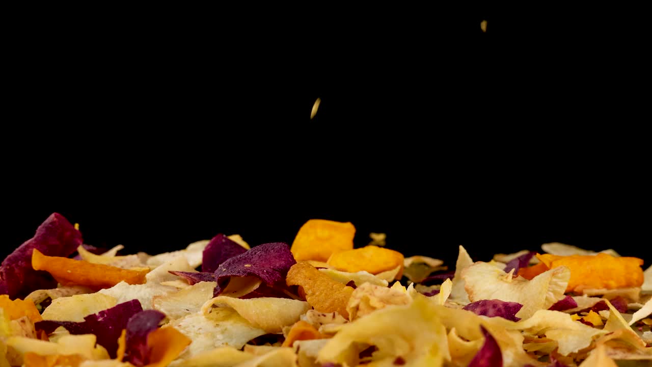 Assorted root vegetable chips fall and scatter on black background, captured in dramatic slow motion