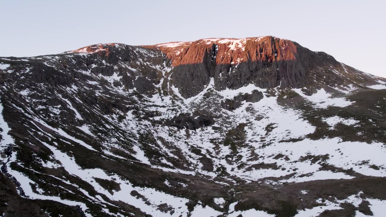 imágenes aéreas de drones que se elevan lentamente para revelar un acantilado montañoso empinado y dramático al amanecer, ubicado en un paisaje salvaje de páramos en la nieve en invierno