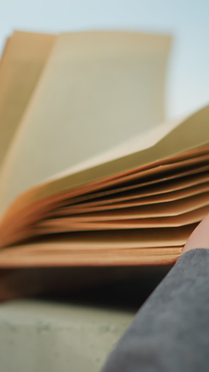 회색 옷을 입은 개인이 오른손으로 열린 책을 들고 서면을 고 바람이 부드럽게 페이지를 가로질러 흐릿한 배경을 가지고 있는 클로즈업 뒷면