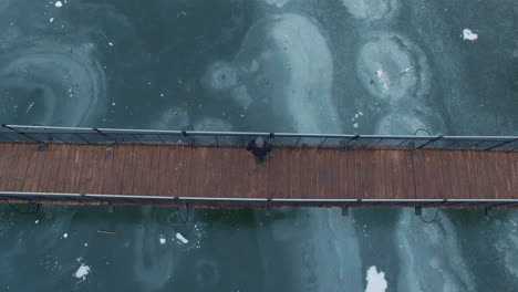 aerial: a man sitting in the middle of a wooden bridge over a frozen lake