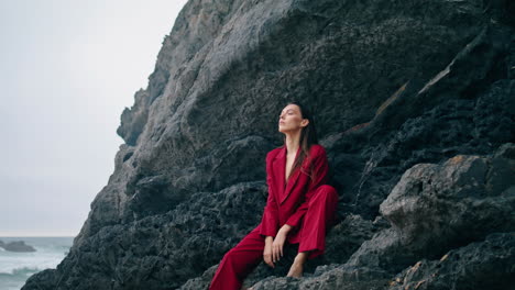 una mujer sentada en una cornisa de roca con un traje rojo. una chica elegante con vista vertical al océano.