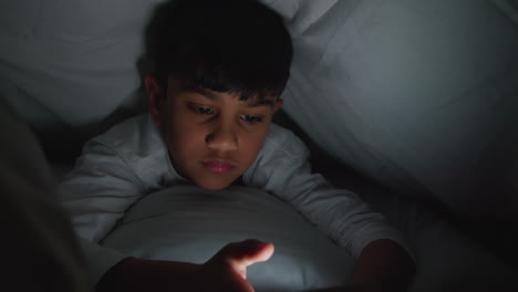 close up of young boy in bedroom at home using mobile phone to text message under covers or duvet at night 3