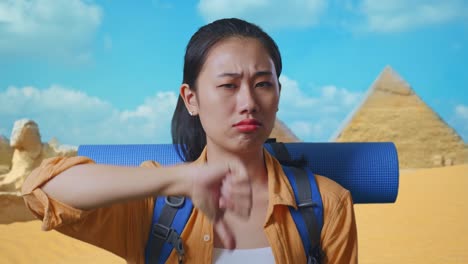 close up of asian female hiker with mountaineering backpack showing thumbs down gesture while traveling in pyramid of giza