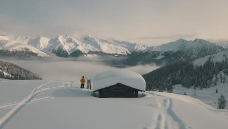 splitboarder stands next to a wooden hut with fresh snow on the roof