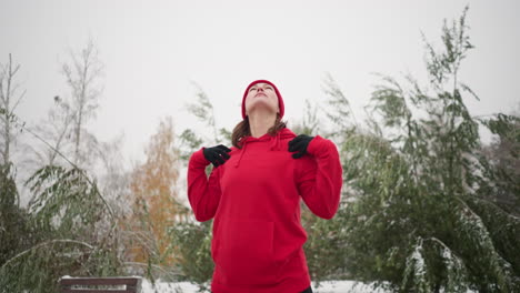 lady performing hand stretch by turning her arm outdoors in winter fitness routine, dressed in athletic wear, snow-covered ground, frosted trees, and bench in serene park setting