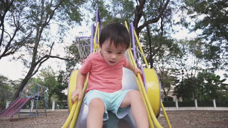 Tapferer-Zweijähriger-Asiatischer-Junge,-Der-Spaß-Daran-Hat,-Zum-Ersten-Mal-Einen-Metallschieber-Hinunterzurutschen-Und-Ein-Wenig-Angst-In-Einem-Outdoor-Park-Zu-Haben