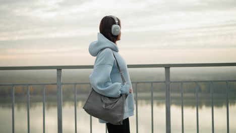 young pretty lady in blue hoodie and black leggings walking with hand holding bag, gazing at river beside her, peaceful outdoor stroll in nature, serene environment, casual and confident
