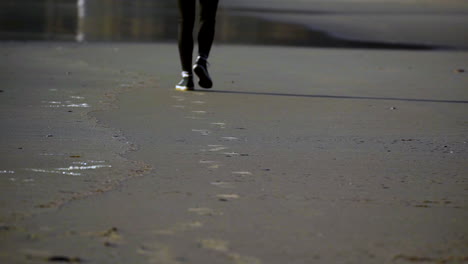 a woman's legs in black pants walking away from the camera leaving footprints in the sand during a sunset at the beach
