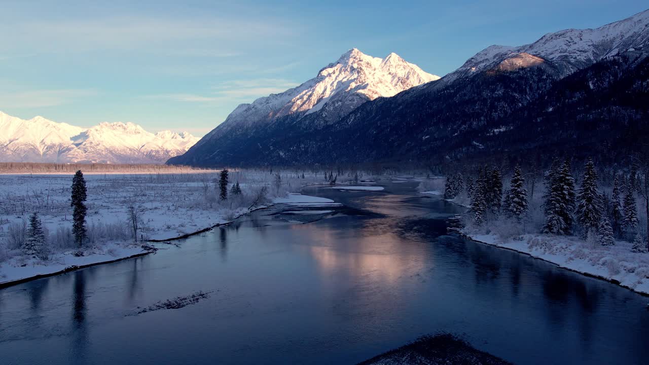 Volo di drone a bassa quota lungo il fiume Knik in profonde ombre. Riflessi del Pioneer Peak innevato visibili nel fiume con il Pioneer Peak in lontananza. 4K, 60FPS.