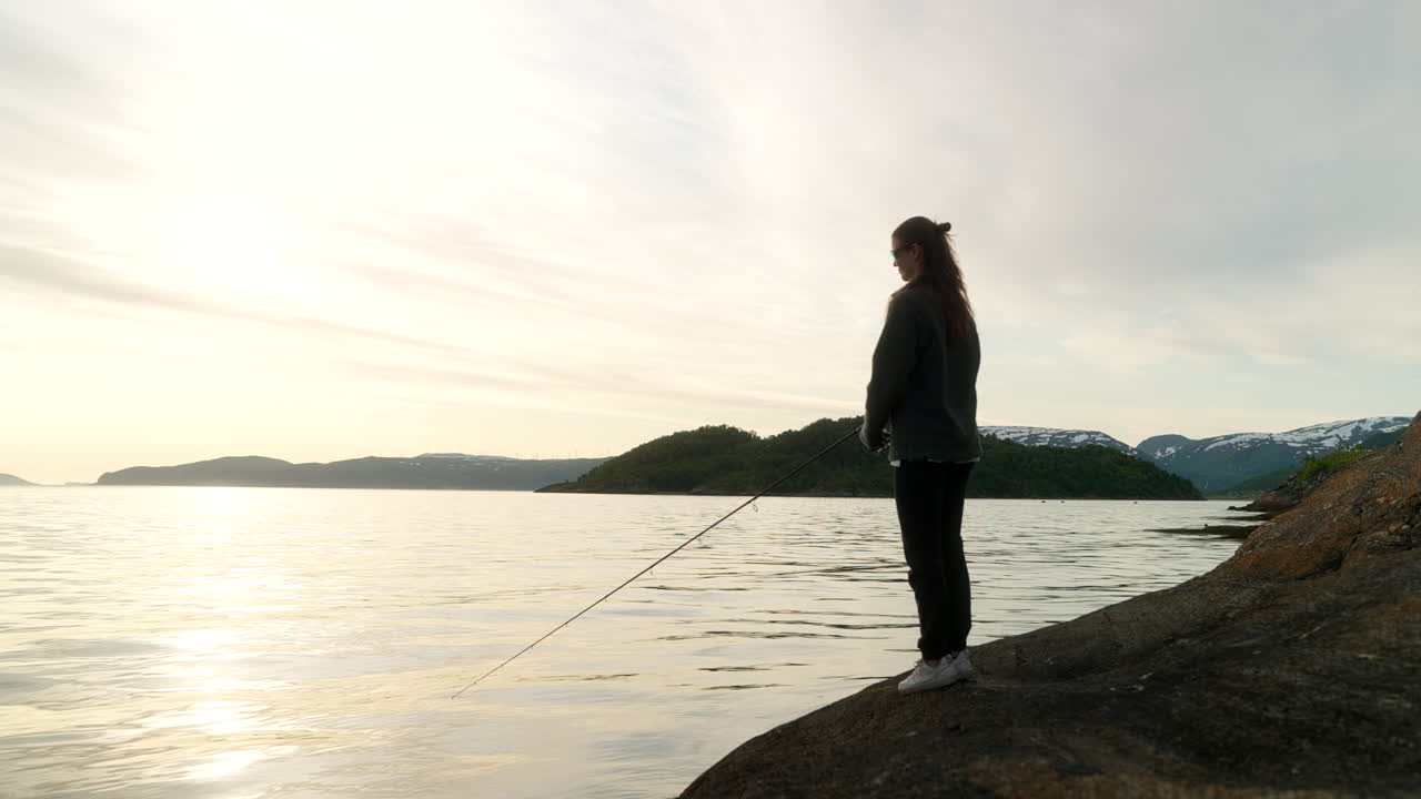 Woman casts pole and fishes in Tromso fjord under soft light, surrounded by still water and hills