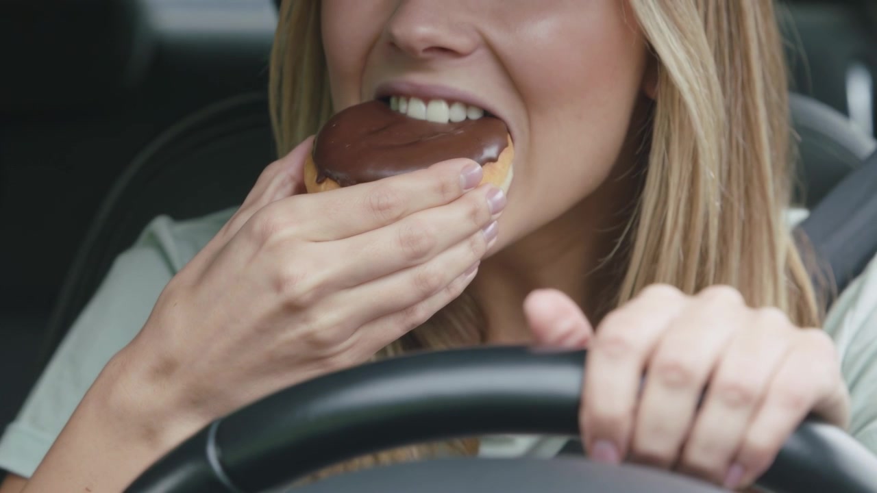 Smiling driver reacting to chocolate-glazed doughnut, bringing it to mouth in car while steering