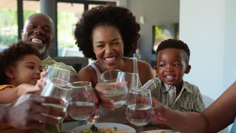 Mehrgenerationenfamilie-Sitzt-Zu-Hause-Am-Tisch-Und-Jubelt-Mit-Wasser-In-Richtung-Kamera