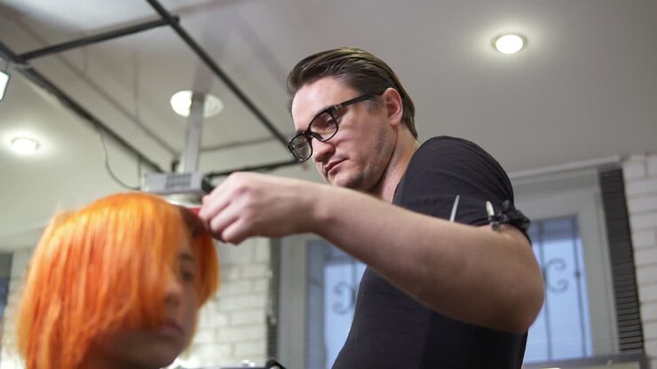 Professional stylish hairdresser in glasses is straightening bright red woman's hair using a hairstraightener in hair salon