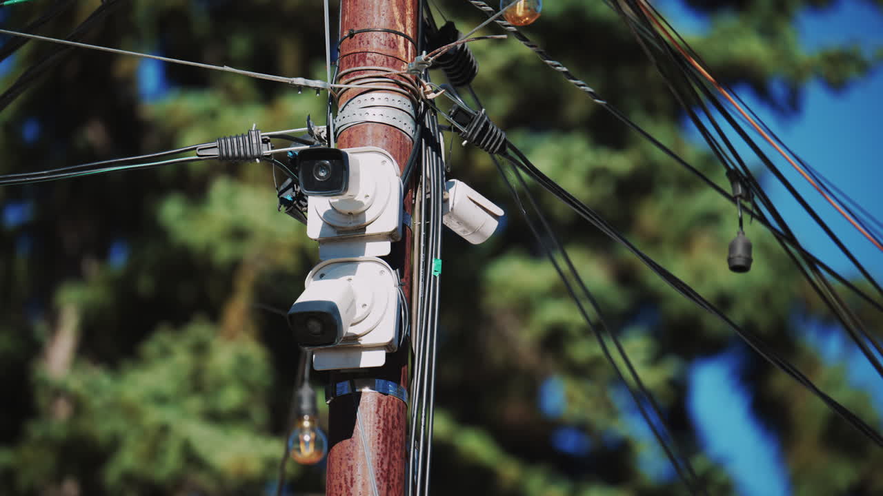 Close up of a utility pole with surveillance cameras, wires, and hanging lights against a blue sky